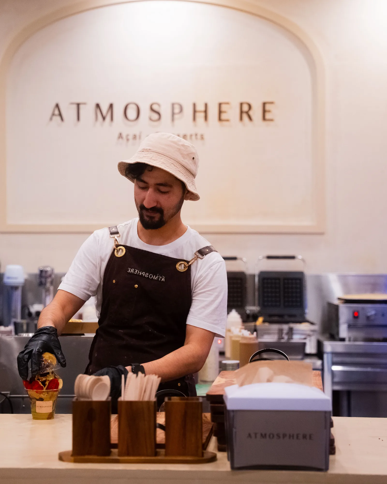 An Atmosphere team member in apron and bucket hat preparing açaí behind the counter